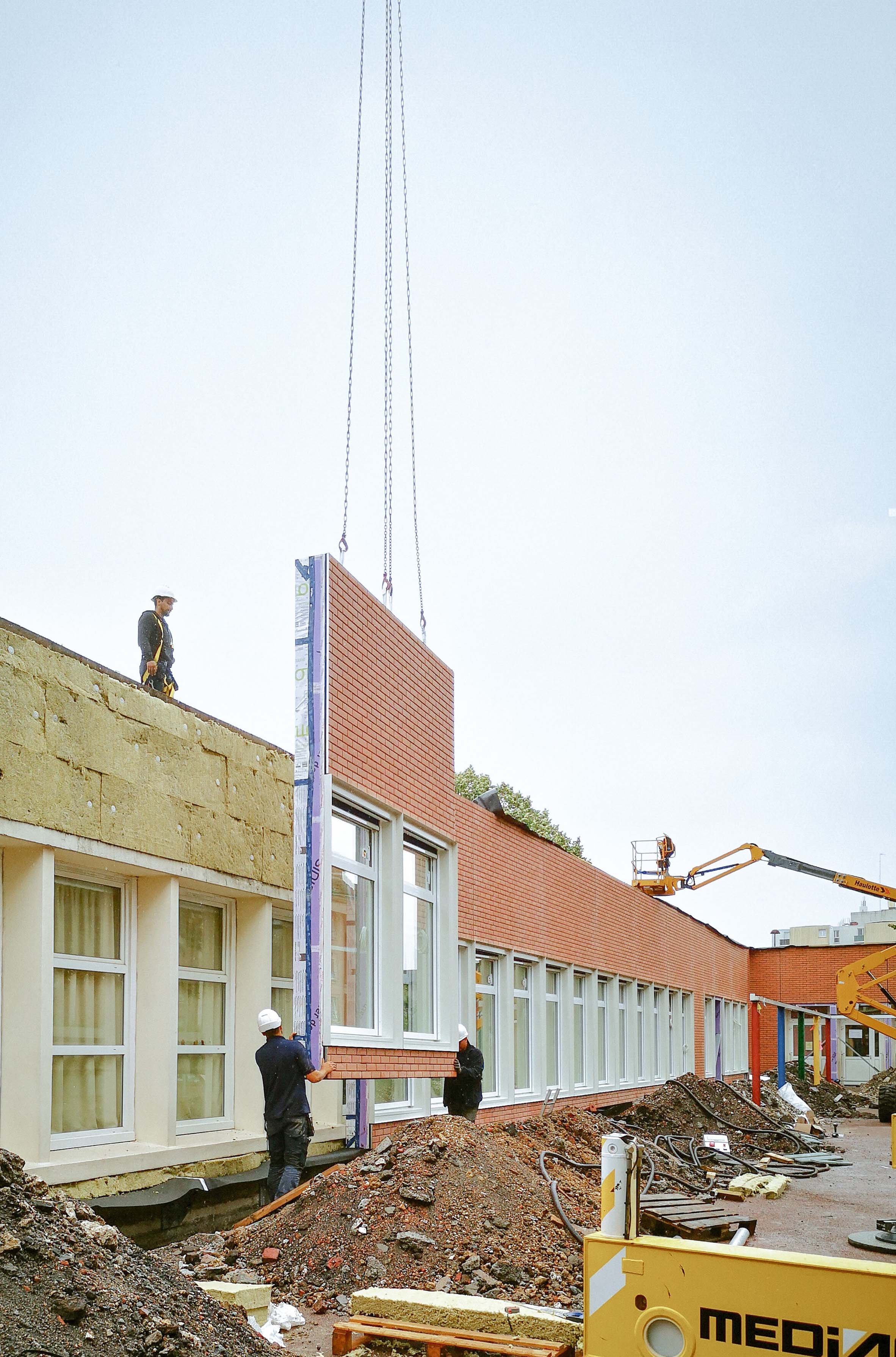 Des ouvriers posent une nouvelle façade vitrée sur un bâtiment scolaire à l'aide d'une grue.