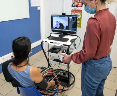 une patiente est assise devant un écran sur lequel apparaît un médecin, elle est assistée par une soignante.