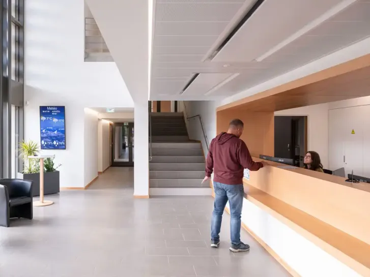 Dans le hall d'accueil d'une mairie, un homme s'adresse à une femme qui est derrière le guichet.