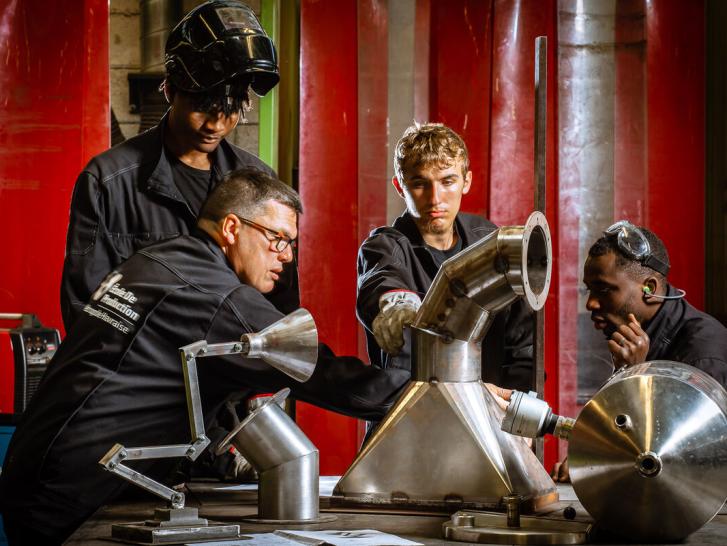 De jeunes apprentis et leur formateur sont en tenue de travail dans un atelier de chaudronnerie.